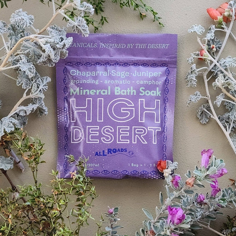 Purple packet of High Desert Mineral Bath Soak surrounded by desert plants and flowers on a beige background